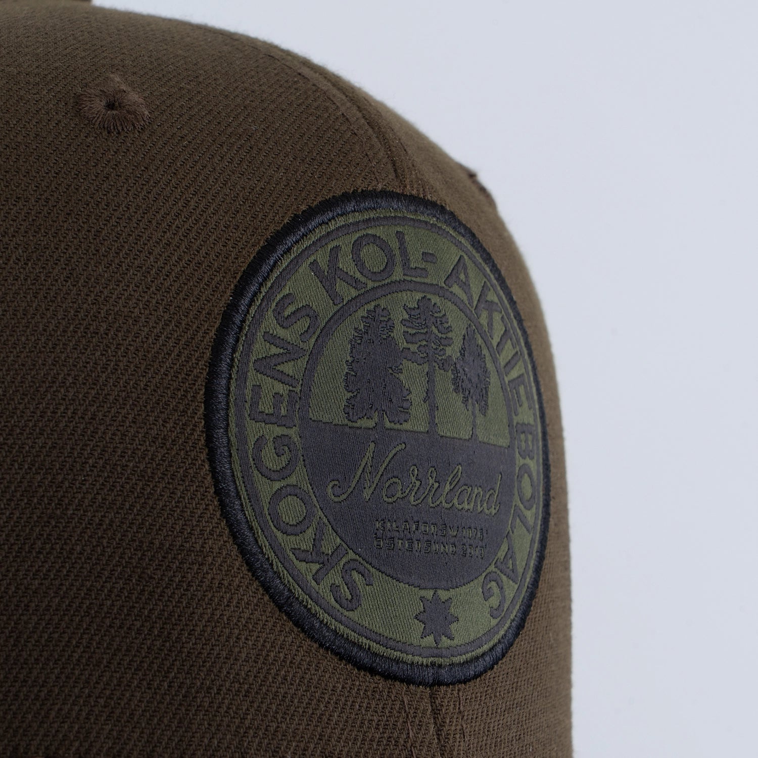 Olivgrön keps med rund broderad patch, text och trädmotiv, fokus på detalj.