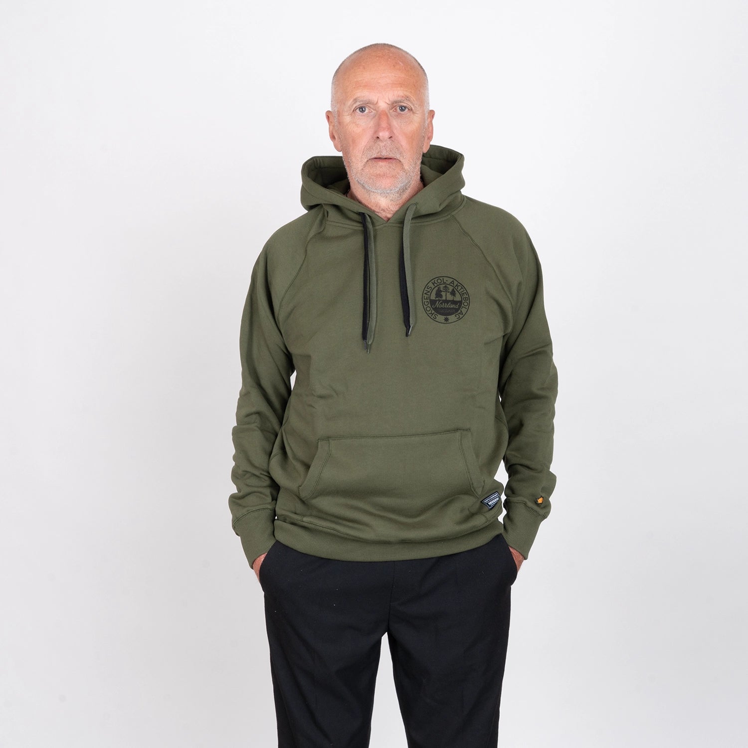 Man i olivgrön hoodie med tryckt logotyp, stående mot vit bakgrund.