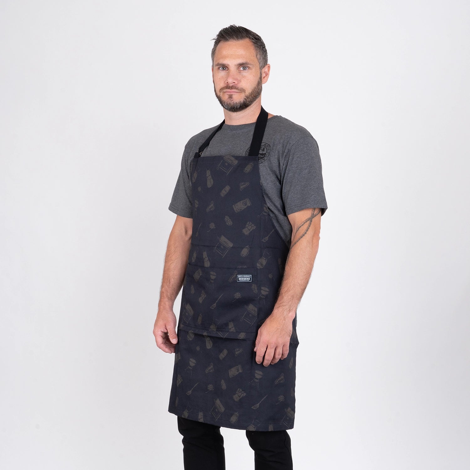 Man i grå t-shirt bär svart grillförkläde, perfekt för utomhusmatlagning och BBQ.