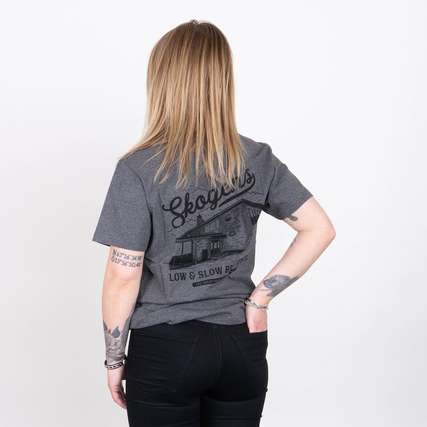 Person i grå t-shirt med grillmotiv, texten 'Skogens Low & Slow BBQ', synliga tatueringar.