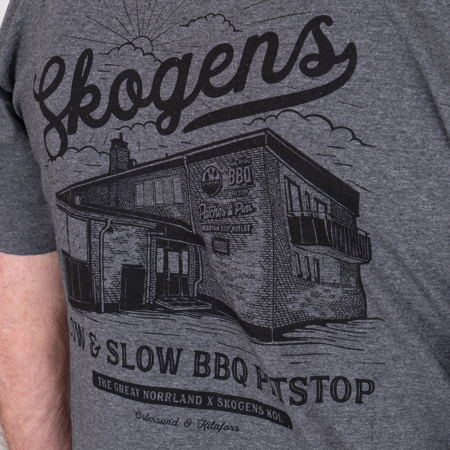 Grå t-shirt med tryck av byggnad, texten Skogens, och grillrelaterade motiv.