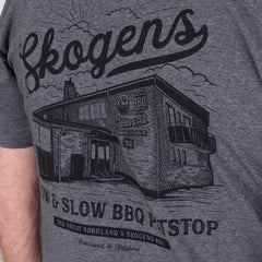Grå t-shirt med tryck av byggnad, texten Skogens, och grillrelaterade motiv.