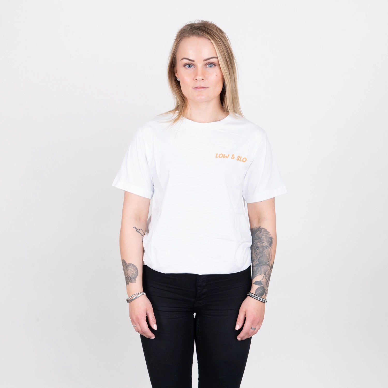 Kvinna i vit t-shirt med orange texten 'LOW & SLO', svart byxa, stående mot vit bakgrund
