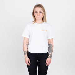 Kvinna i vit t-shirt med orange texten 'LOW & SLO', svart byxa, stående mot vit bakgrund