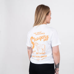 Kvinna i vit t-shirt med orange tryck, motiv av ko och text om Norrland och grillning.