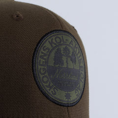 Olivgrön keps med rund broderad patch, text och trädmotiv, fokus på detalj.