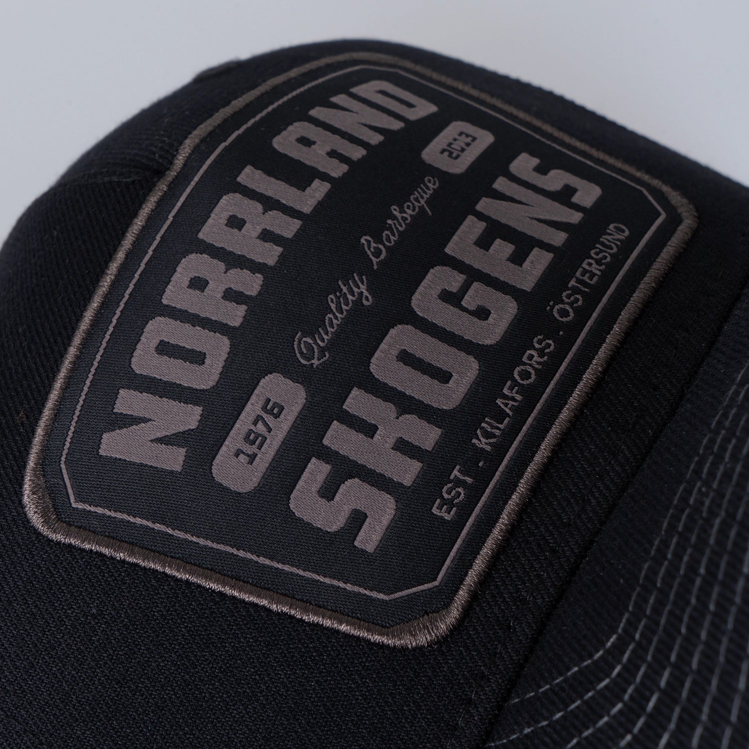 Svart truckerkeps med brodyrpatch, text NORRLAND SKOGENS, grill och outdoor-accessoar.