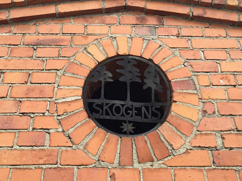 Rund fönsterdekor i smide med texten SKOGENS och trädmotiv i tegelvägg